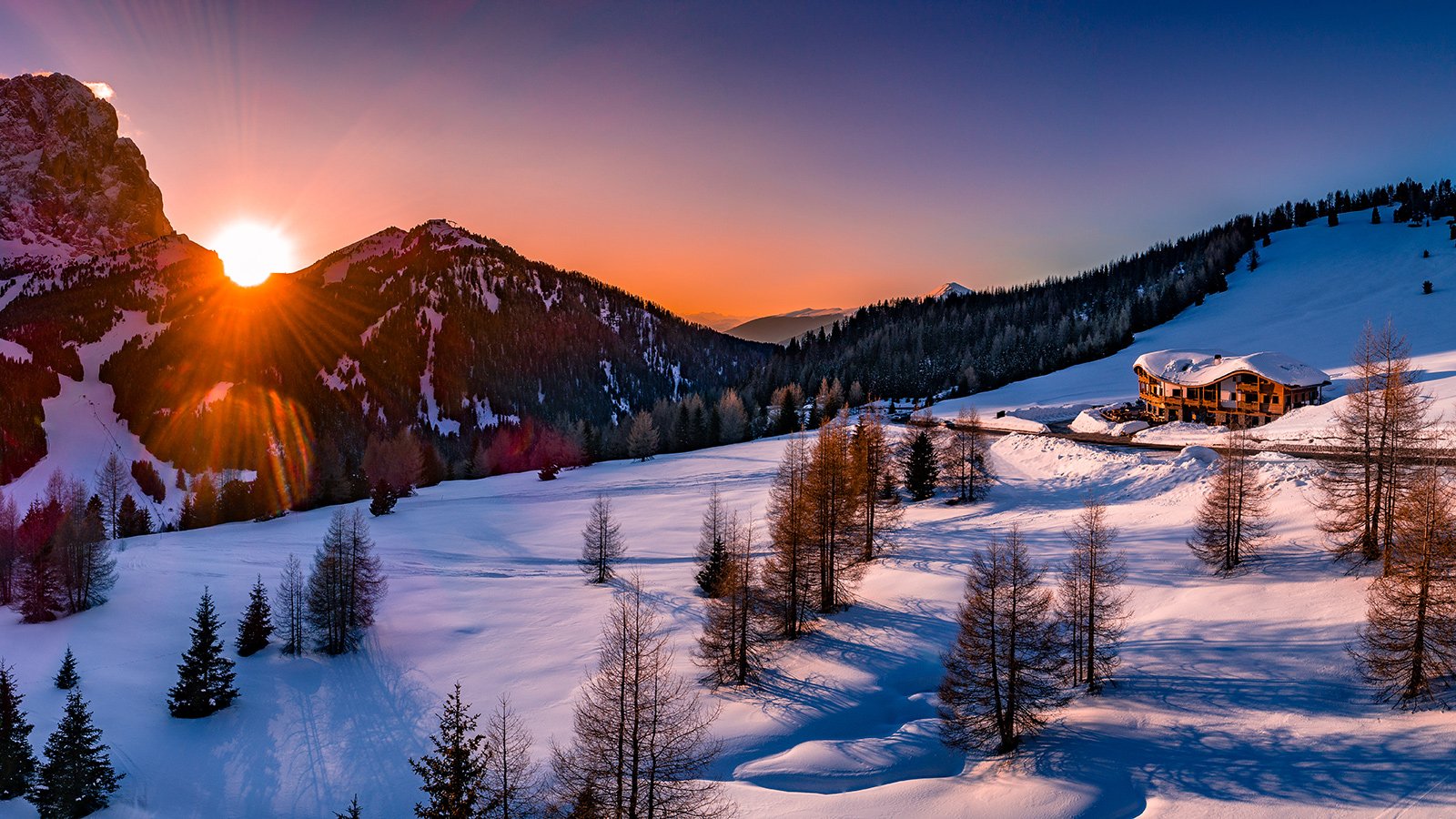 Winter landscape with Chalet Gerard on the snow as the sun sets behind the mountains, creating a colorful sky