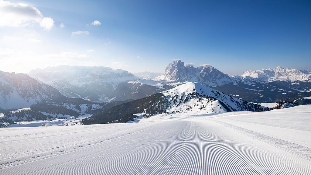 A perfectly groomed ski slope in the morning, with the typical corduroy lines on the snow, and a panoramic view of the Dolomite peaks